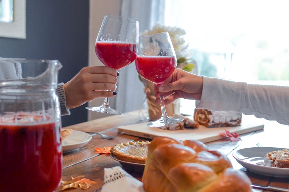 Two people clink glasses with red beverages in a cozy indoor setting surrounded by pastries and pie.