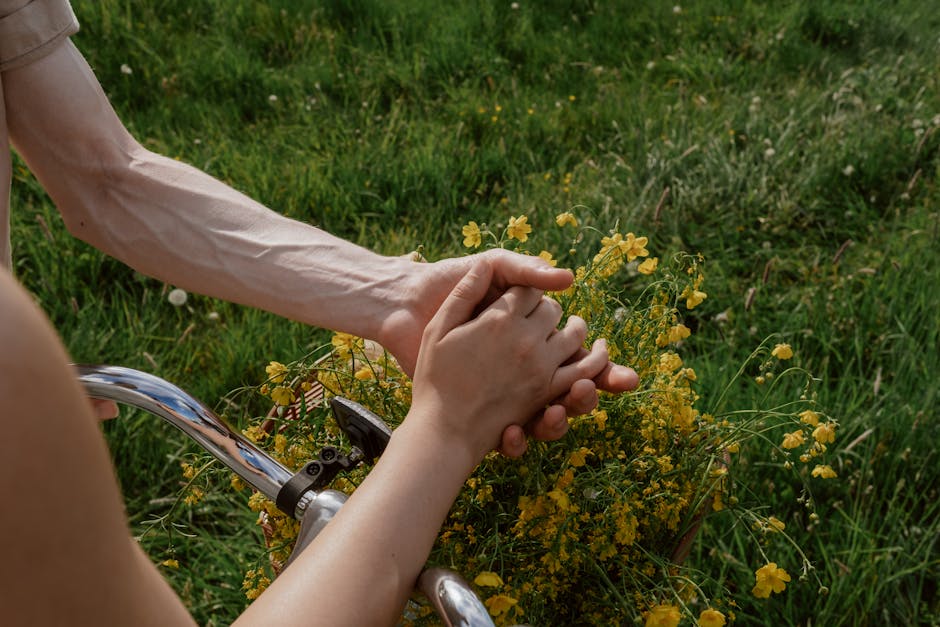 A close-up of a couple holding hands over bicycle handlebars with yellow flowers in spring.
