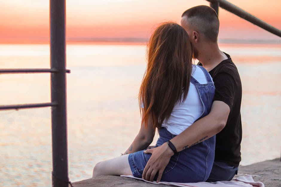 A couple sits on a pier, embracing as they watch a beautiful sunset over the sea.