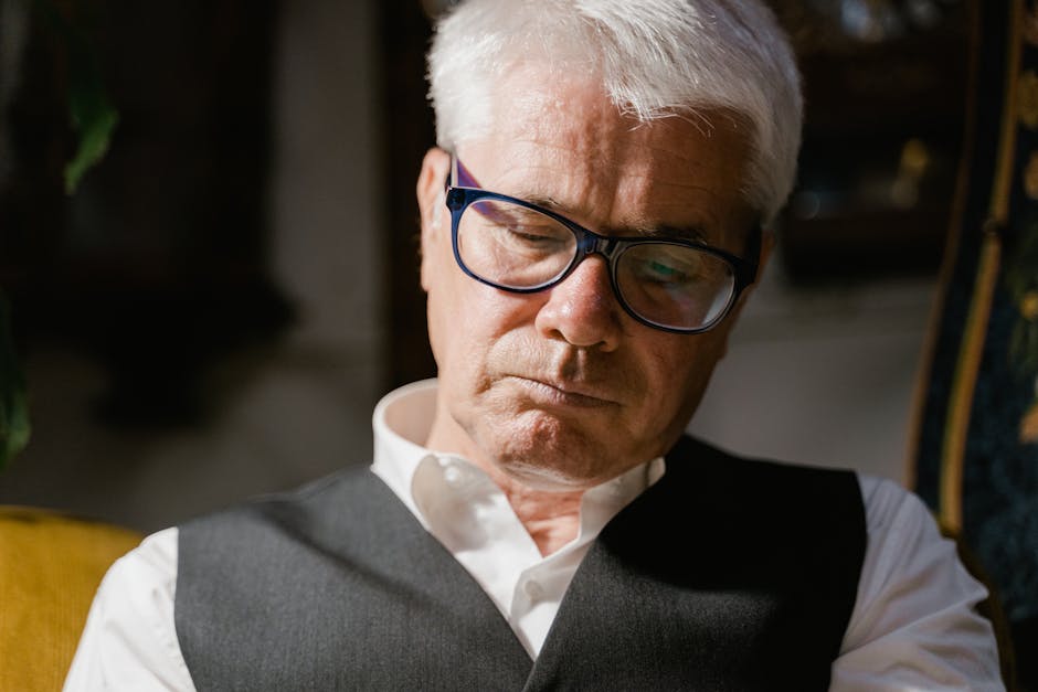 Thoughtful elderly man with glasses and grey hair sitting indoors.