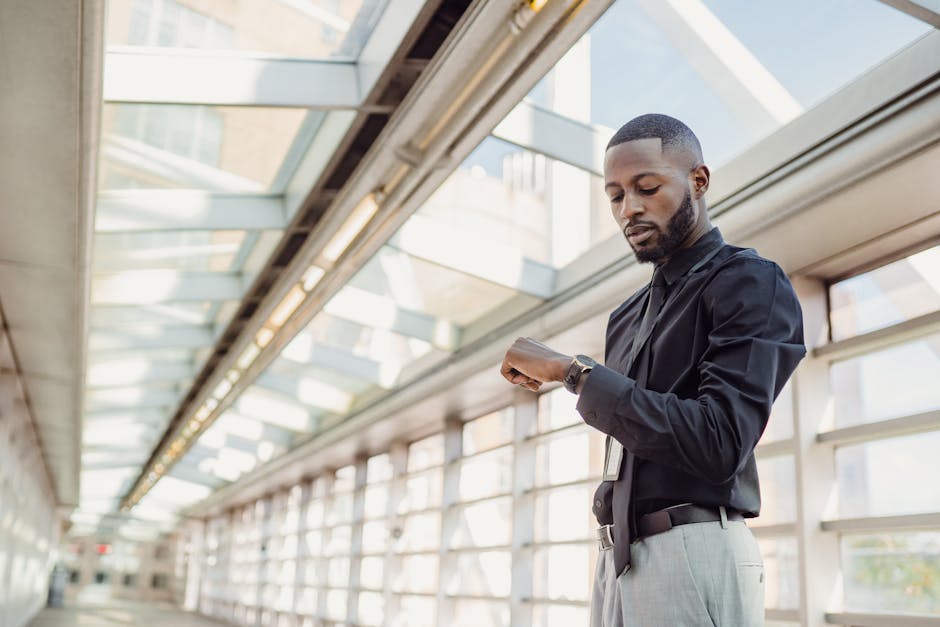 African American man in shirt checks wristwatch, standing in vibrant modern glass corridor.