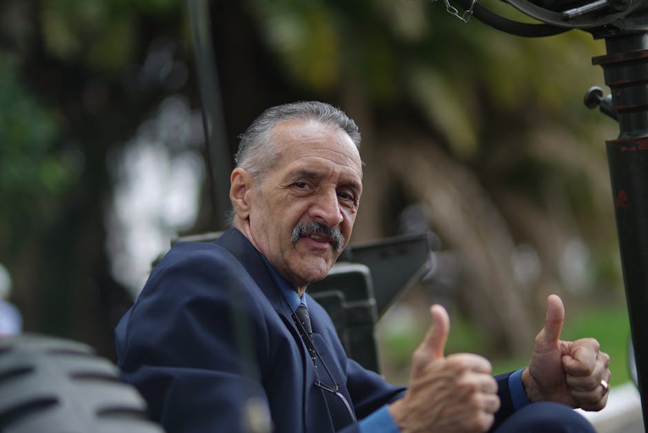 Elderly man in a suit gives a thumbs up gesture, sitting outdoors with trees in the background.