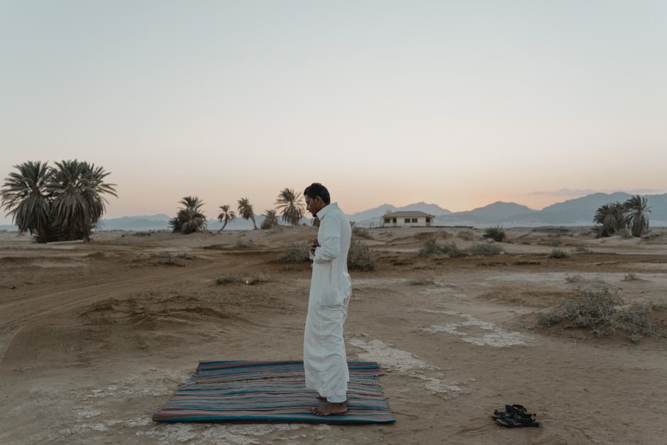 A man stands on a prayer mat in the desert, performing a religious ritual at sunrise.