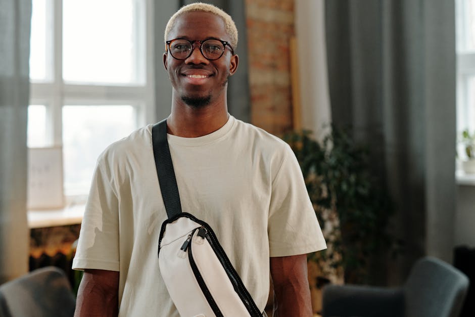Casually dressed man with eyeglasses and bag smiling indoors against a bright window.