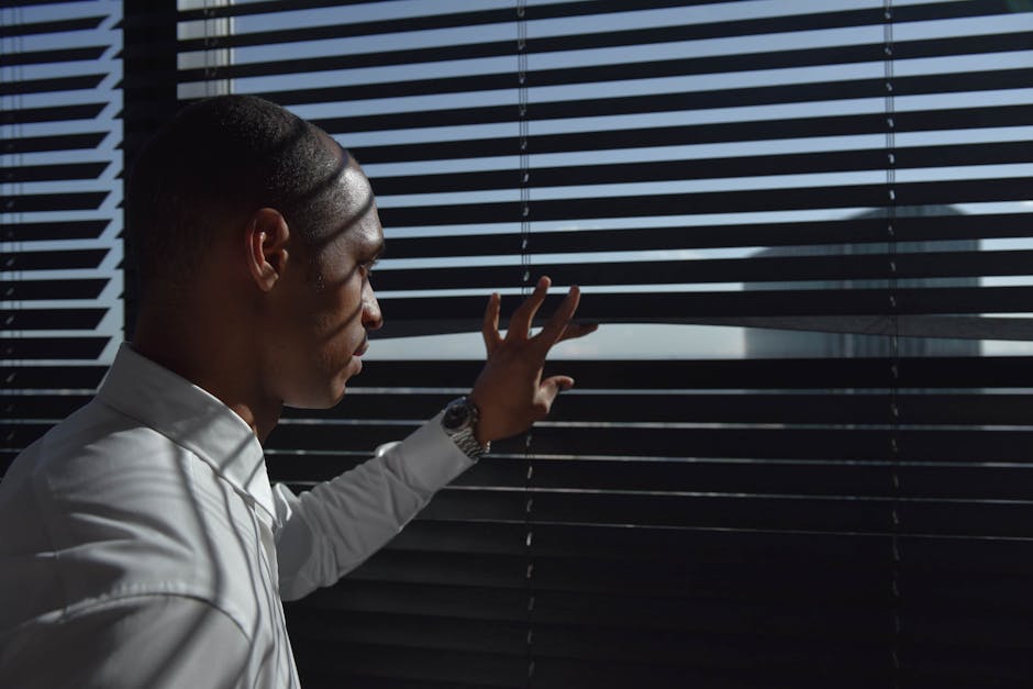A man looks through window blinds in an office setting, creating a mysterious ambiance.