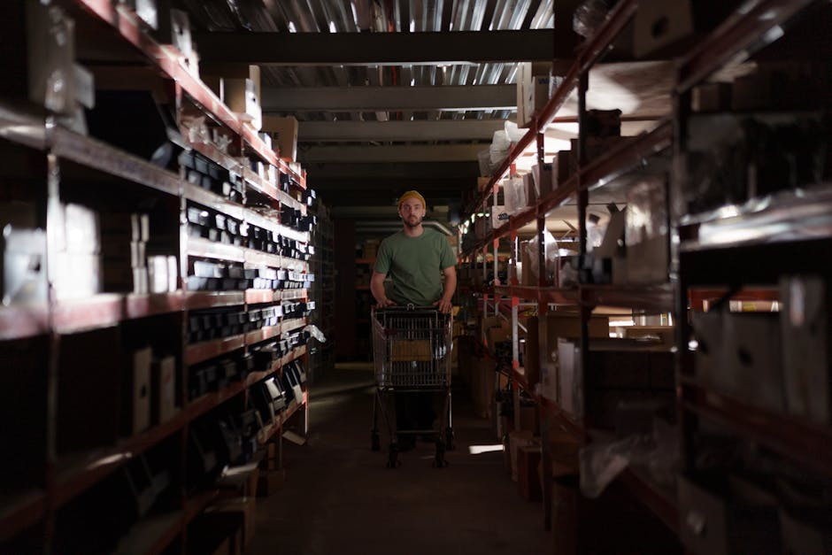 A man wearing a beanie pushes a cart through a dimly lit warehouse aisle.