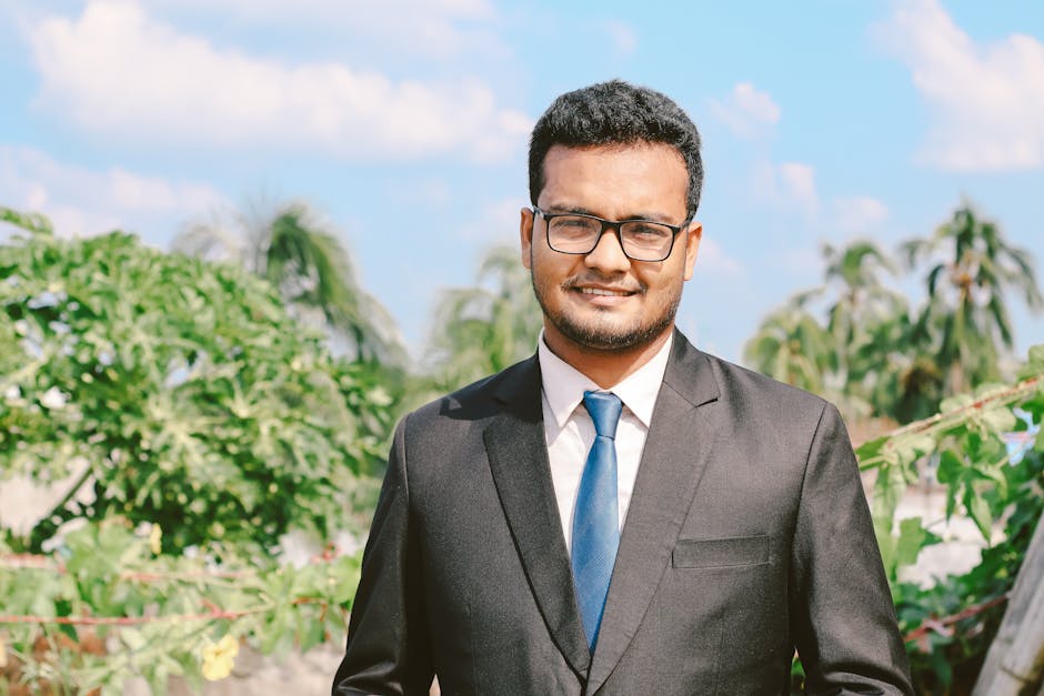 Portrait of a smiling man in a suit and eyeglasses, exuding confidence outdoors with a clear blue sky.